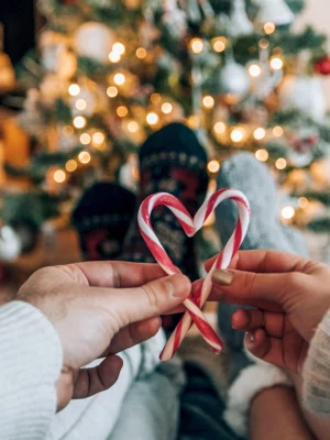 A selective focus shot of a couple in knitted socks holding heart-shaped candy canes with a glowing bright Christmas tree in the background